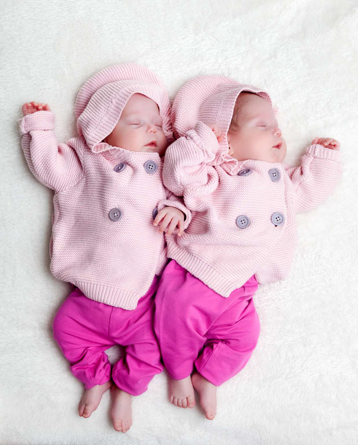 Newborn twins sisters sleeping on white fur, wearing pink sweaters